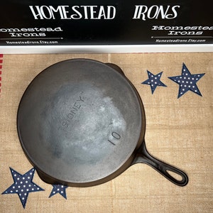 May include: A 10-inch cast iron skillet with the word "SIDNEY" embossed on the bottom. The skillet is sitting on a burlap surface with red and white stripes.