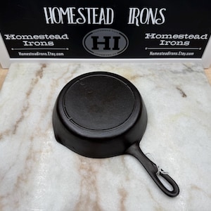 May include: A black cast iron skillet with a single handle. The skillet is round with a slightly sloped side. The handle has a hole for hanging. The background includes a sign that says "Homestead Irons" and the website address.
