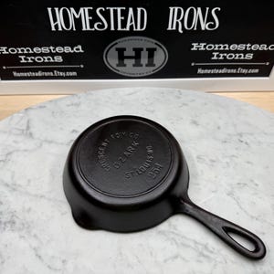 May include: A black cast iron skillet with a handle, resting on a marble surface. The skillet has the text "CRESCENT FDY CO OZARK ST. LOUIS, MO 254" embossed on the bottom. The sign in the background reads "HOMESTEAD IRONS" and "Homesteadirons.Etsy.com".