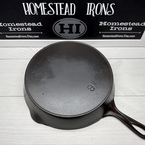 May include: A black cast iron skillet with a long handle and a circular cooking surface. The number "9A" is visible on the bottom. The brand name "SIDNEY" is embossed on the side. The sign above reads "HOMESTEAD IRONS".