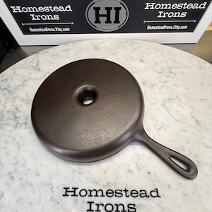 May include: A dark brown cast iron skillet lid with a handle and a hole in the center. The lid is round and sits on a white and gray surface. The text "Homestead Irons" and "Homesteadirons.Etsy.com" are visible.