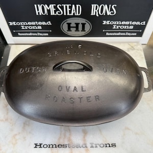 May include: A dark gray cast iron oval roaster with a lid featuring the embossed text No 5 GRISWOLD DUTCH OVEN OVAL ROASTER. The lid has a central handle and two side handles. The background sign displays the text HOMESTEAD IRONS and HI.