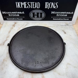 May include: A round, black cast iron skillet with a textured surface. The skillet has a raised edge and two small handles. The number "12 A" is stamped on the surface. The sign above reads "Homestead Irons".