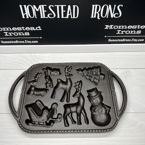 May include: A black cast iron baking pan with a Christmas theme. The pan has eight molds, including a snowman, a Christmas tree, and reindeer. The pan is made by Homestead Irons.