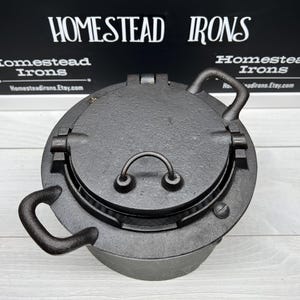 May include: A black cast iron waffle iron with a hinged lid and two handles. The iron has a waffle pattern on the inside of the lid. The iron is sitting on a white wooden surface.