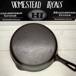 May include: A dark brown cast iron skillet with a long handle. The skillet has the text "Wagner Ware Sidney -0-" and the number "1129" embossed on it. The background includes a sign that says "Homestead Irons" and "Homesteadirons.Etsy.com".
