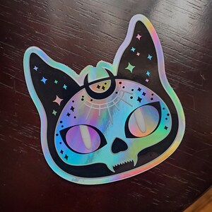 May include: A holographic sticker of a black cat with a crescent moon on its head. The cat's eyes are iridescent blue and the sticker has a glittery finish.