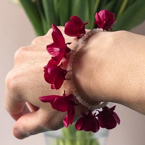 May include: A floral bracelet featuring deep red flower accents strung on a light-colored beaded strand. The bracelet is worn on a wrist, with a bouquet of pink and white tulips in the background. The flowers are in a clear glass vase.