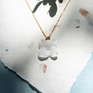 Peut inclure: Un collier doré délicat avec un pendentif en forme de fleur blanche. Le pendentif est une fleur pressée à quatre pétales avec un centre rose pâle. Le collier repose sur une surface texturée blanc cassé, avec des ombres et de la lumière.