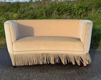 May include: A light beige curved sofa with a fringe trim. The sofa is sitting on a black tarmac surface.