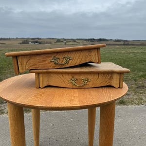 May include: Two wooden boxes with brass handles are stacked on a round wooden table with four legs. The boxes and table are made of light-colored wood. The background shows a field and a cloudy sky.