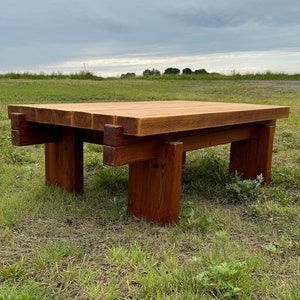 Könnte beinhalten: Ein rustikaler Holz-Couchtisch mit einer rechteckigen Tischplatte und vier quadratischen Beinen. Der Tisch ist aus dunkelbraunem Holz gefertigt und hat eine natürliche Oberfläche. Der Tisch steht auf einer grasbewachsenen Wiese.