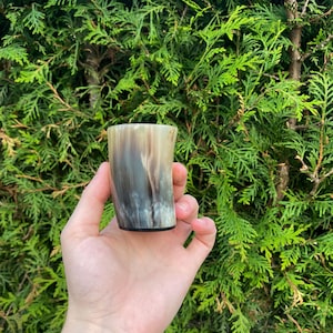Peut inclure: Une petite tasse à boire artisanale en corne naturelle. La tasse présente un motif unique de teintes marron, grises et blanches. La tasse est tenue dans une main devant un fond de feuillage vert.