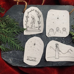 May include: A slate-colored decorative piece with four hand-drawn images depicting a nativity scene. The images are in black ink on a textured, off-white paper. The piece is decorated with evergreen branches and a red background.