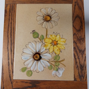 May include: A framed ceramic tile featuring a floral design. The tile depicts three white and yellow flowers with green leaves on a beige background. The frame is made of dark wood.
