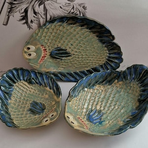 May include: Three ceramic fish-shaped serving dishes in shades of blue, green, and beige. The dishes have raised scales and googly eyes.