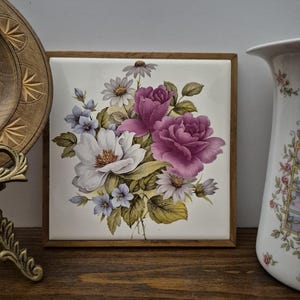May include: A square floral tile with a wooden frame. The tile features a bouquet of white, purple, and blue flowers with green leaves. The tile is set on a wooden surface.