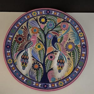 May include: A round, hand-painted decorative plate featuring a tree with colorful birds and flowers. The plate has a pink and purple border with floral accents. The birds are pink and blue, with detailed feather patterns.