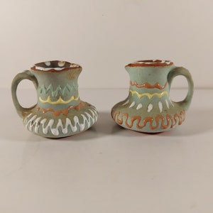 May include: Two small, hand-painted ceramic jugs with a light blue glaze and colourful geometric designs. The jugs have a rounded body and a small handle.