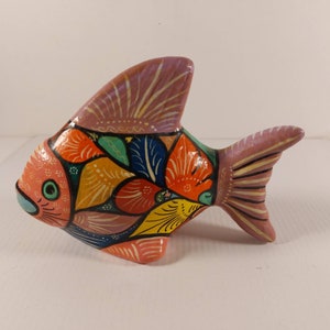 May include: A colourful, hand-painted ceramic fish ornament. The fish has a coral-coloured body, a lavender dorsal fin, and a tail. The body is decorated with orange, yellow, blue, and green patterns. The fish has a green eye and black details.