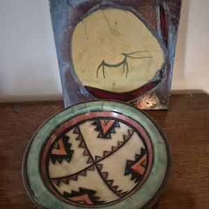 May include: Two ceramic pieces: a square, abstract painting with a yellow circle and a deer-like figure, and a round bowl with a geometric design in green, pink, and orange. The bowl has a light green rim.