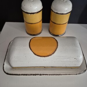 May include: A ceramic butter dish and two salt and pepper shakers. The butter dish is rectangular with a white base, a brown rim, and a large orange circle in the center. The shakers are cylindrical with white tops, yellow and orange sections.