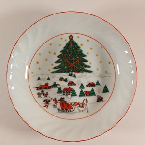 May include: A white ceramic Christmas plate with a red rim. The plate features a winter scene with a green Christmas tree, red and white houses, and a red sleigh pulled by a white horse.