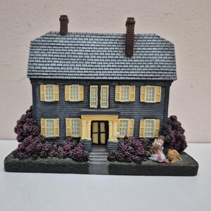 May include: A miniature two-story house figurine with dark blue siding, a gray shingled roof, and two brown chimneys. The house features yellow shutters, a central yellow doorway, and dark purple bushes at the base. A small figure and a dog sit outside.