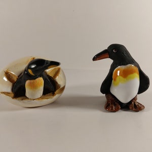 May include: Two ceramic penguin figurines. One is hatching from an egg, the other stands upright. Both have black bodies, white bellies, and yellow and orange details. The figurines are on a white surface.