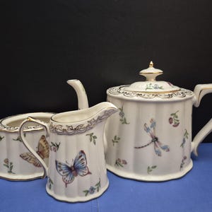 May include: A white porcelain tea set featuring a teapot, a creamer, and a sugar bowl. Each piece has a fluted body, gold-trimmed edges, and a delicate pattern of colorful butterflies, dragonflies, and small floral sprigs scattered across the surface.