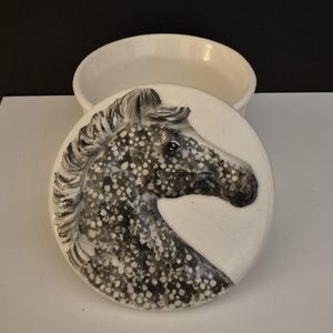 May include: A white ceramic trinket box with a lid featuring a detailed horse head design. The horse head is rendered in shades of grey and black with a speckled pattern. The box is round and has a simple, elegant design.