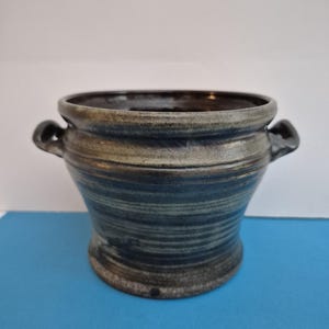 May include: A dark gray ceramic pot with two handles. The pot has a striped pattern and a slightly flared rim.