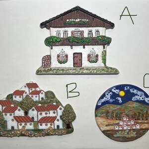 May include: Three ceramic decorative pieces featuring miniature village scenes. One is a house with a brown roof and white walls, another depicts a cluster of houses on a hill, and the third is a round piece with a night scene.