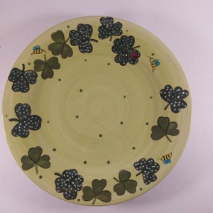 May include: A green ceramic dinner plate with a white rim and a design of black shamrocks with white dots. The shamrocks are arranged around the edge of the plate, and there are also small black dots scattered across the plate. There are also small images of bees and ladybugs on the plate.
