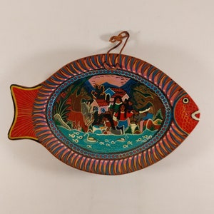 Peut inclure: Une suspension murale en bois en forme de poisson colorée avec une scène peinte d'un village avec des personnes et des animaux. Le poisson est rouge, orange et bleu avec un design détaillé.