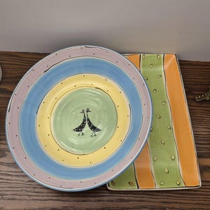 May include: A pair of decorative ceramic plates. A large, round bowl features concentric rings of blue, yellow, and lilac, with a green centre and two bird figures. The rectangular serving dish has orange, green, and yellow stripes.