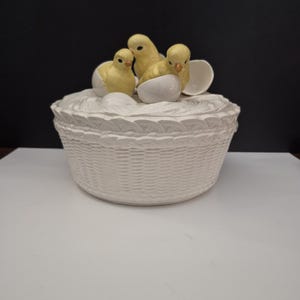 May include: A white ceramic basket-shaped container with a textured woven design. Three yellow chicks are perched on top, emerging from white eggshells. The chicks have orange beaks and are a charming Easter-themed decoration.