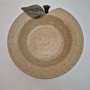 May include: A beige ceramic bowl shaped like an apple with a green leaf and brown stem. The bowl has a speckled finish.