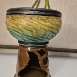 May include: A ceramic candle holder with a distinctive design. The top bowl has a blend of yellow, green, and blue colours, while the base is a warm brown with decorative cutouts. The rim is black. This decorative piece is suitable for home decor.