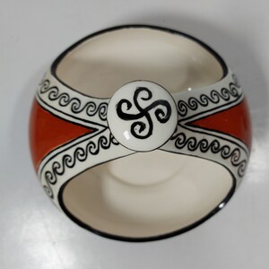 May include: A white ceramic bowl with a red band and black wavy lines. The bowl has a handle with a black and white design.
