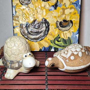 May include: Two ceramic turtle ornaments. One is light beige with a textured shell and a white face. The other is brown with white floral designs. A painting of sunflowers in yellow, blue, and black is in the background.