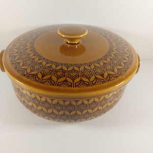 May include: A brown lidded casserole dish with a repeating geometric pattern. The pattern is a darker shade of brown. The dish has two small handles and a knob on the lid. The dish is suitable for cooking.