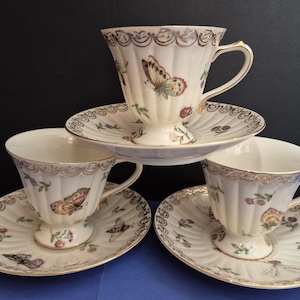 May include: Three cream-colored porcelain teacups and matching saucers featuring a ribbed texture, gold-trimmed scalloped edges, and colorful butterfly and floral motifs. Each cup has a pedestal base and a curved handle with gold accents.