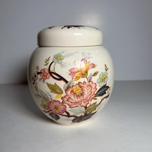 May include: A cream-colored ceramic jar with a lid, adorned with a floral design in shades of pink, yellow, blue, and green. The jar has a rounded shape and a matching lid, creating a decorative storage container.