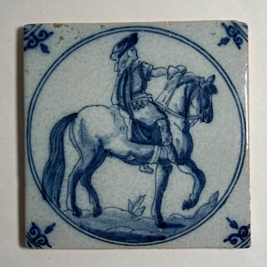 May include: Square ceramic tile with a hand-painted design of a person on horseback in shades of blue. The rider is wearing a hat and period clothing. The tile has a circular border and decorative corner accents.