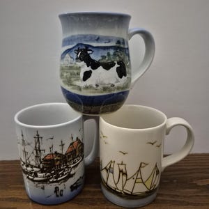 May include: Three ceramic mugs with different designs. One mug features a black and white cow in a landscape scene. Another mug has a nautical theme with a harbor scene. The third mug has a sailboat design.
