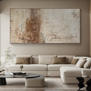 May include: Abstract painting with brown and white tones. The painting is hanging on a wall above a white sectional sofa with brown throw pillows.