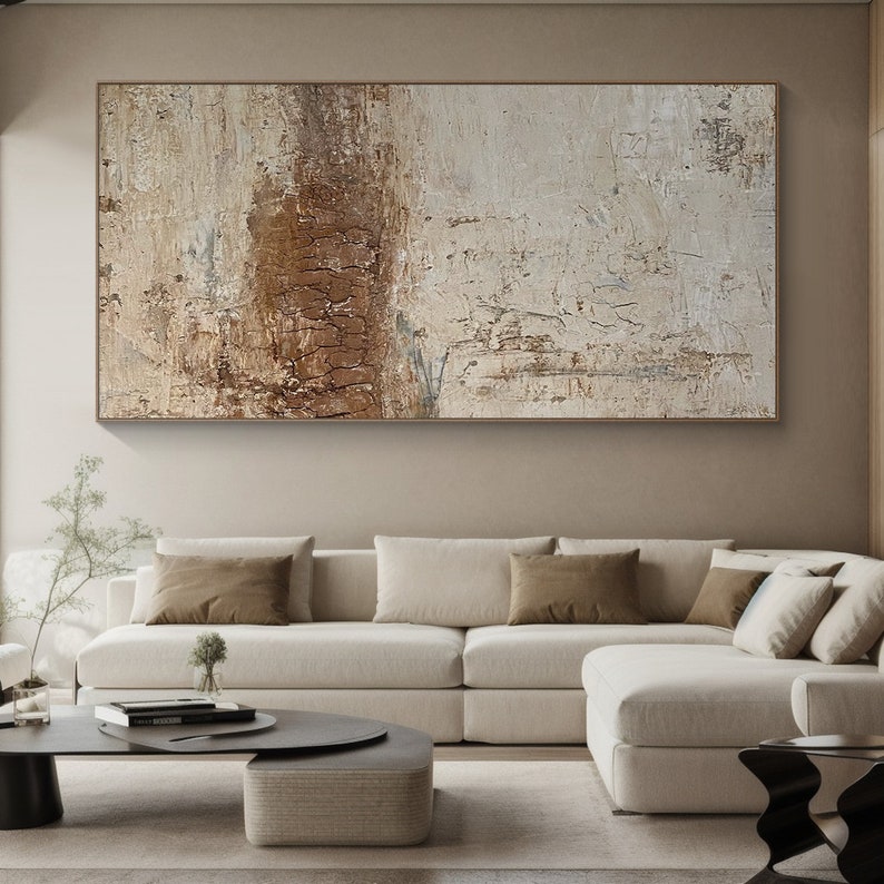 May include: Abstract painting with brown and white tones. The painting is hanging on a wall above a white sectional sofa with brown throw pillows.