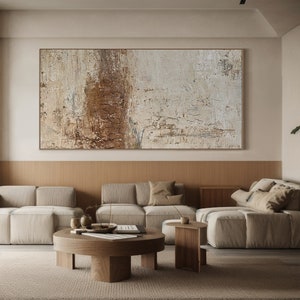 May include: A living room with a large abstract painting in beige and brown tones hanging on the wall above a wooden panel. The room is furnished with a beige sectional sofa, a coffee table, and two side tables.