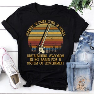 May include: Black t-shirt featuring a vintage-style sunset graphic with a sword and the text "Strange Women Lying in Ponds Distributing Swords is No Basis for a System of Government."